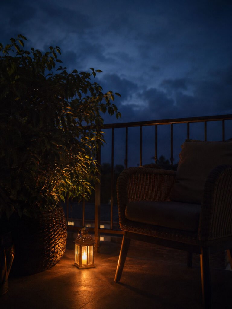 Small warm lantern glowing on the floor beside a balcony chair at dusk, with soft light on nearby plants and a dark sky above