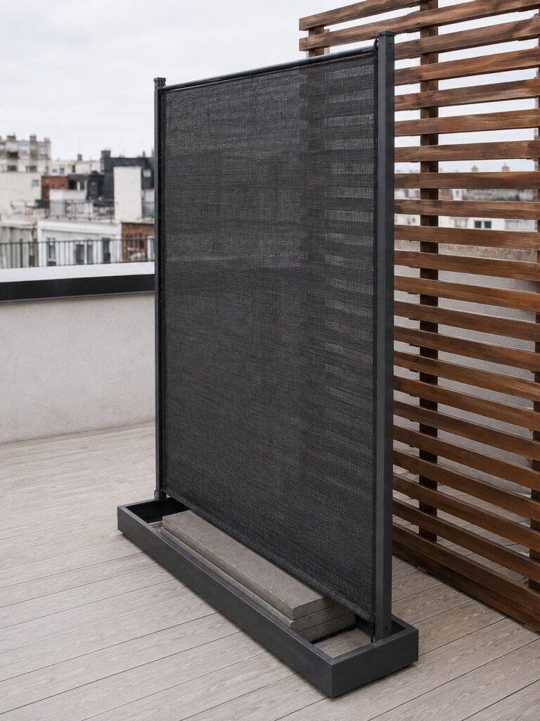 A dark mesh privacy panel in a low frame on a rooftop with paving stones stacked inside the base
