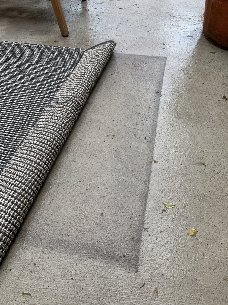 A charcoal flat woven outdoor rug lifted at one edge on a concrete patio, showing a darker damp outline on the slab underneath