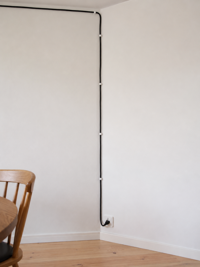 A pendant cord running along the ceiling line and down a wall corner, held by small clips, ending at a wall outlet above the baseboard